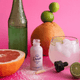Bottle of 'Drops' with a dropper, citrus fruits, and a pink drink on a pink background